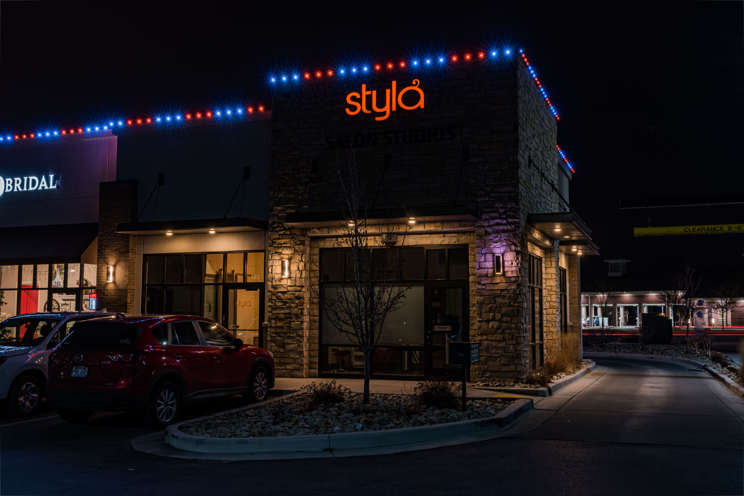 A salon named stylā with orange neon signage is illuminated at night. The building has stone walls and is decorated with blue and red string lights along the roof. A red SUV is parked outside.