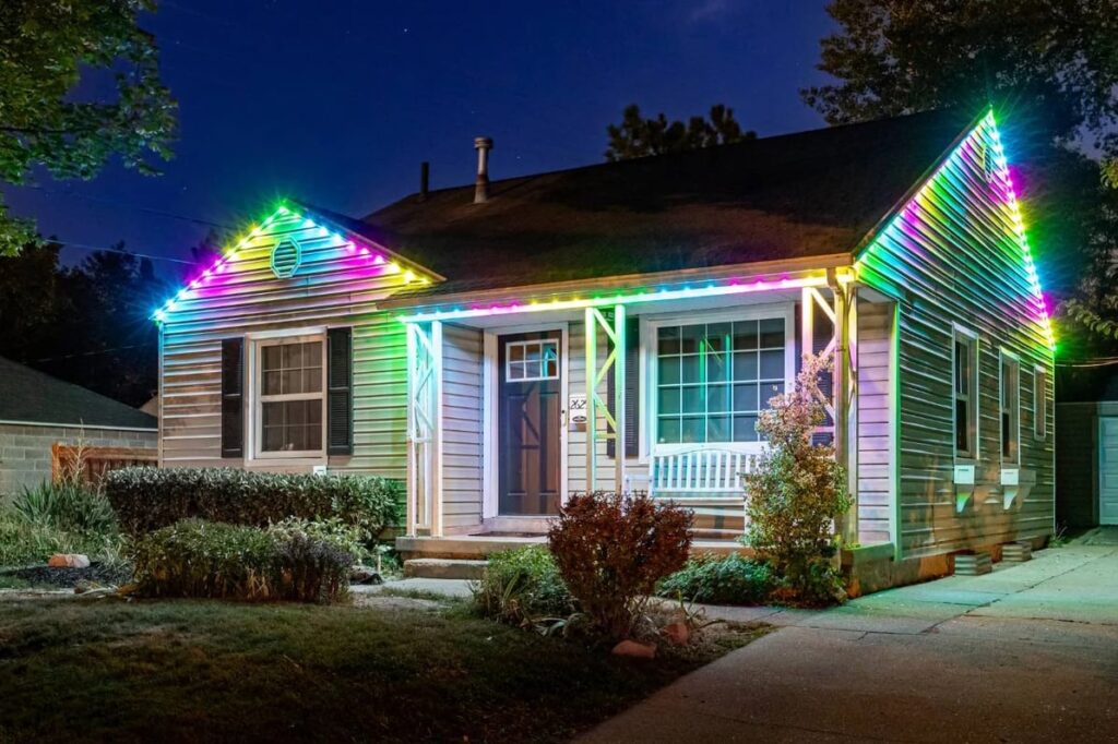 A small house at night with its roofline decorated by colorful, bright LED lights. The yard has bushes and a white bench sits by the front door. The house is well-lit and inviting.