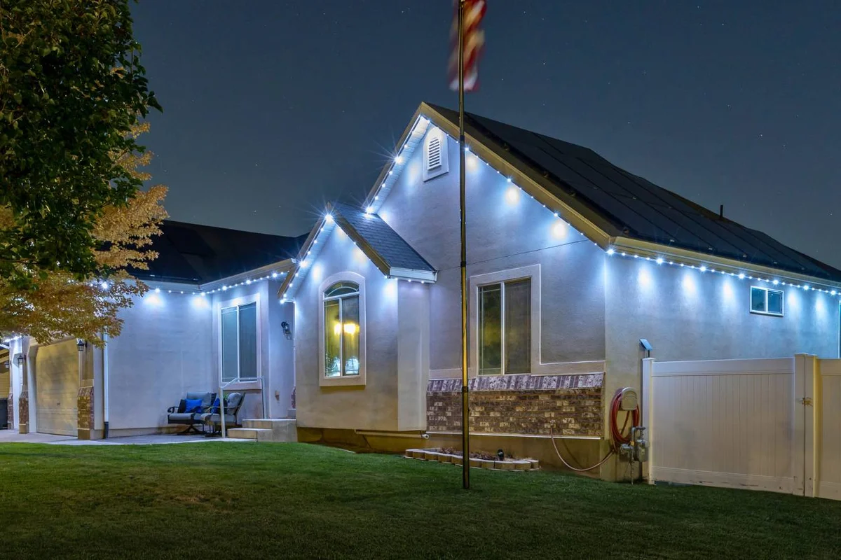 A single-story house at night with white exterior walls, outdoor string lights along the roofline, a flagpole in front, a manicured lawn, and a tree partially visible on the left.
