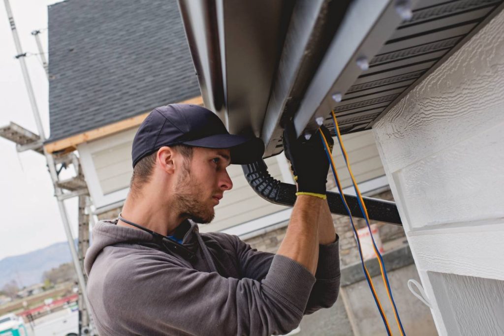 A man wearing a cap and gloves installs or repairs a rain gutter along the roof edge of a house, concentrating on his work with tools and materials visible.