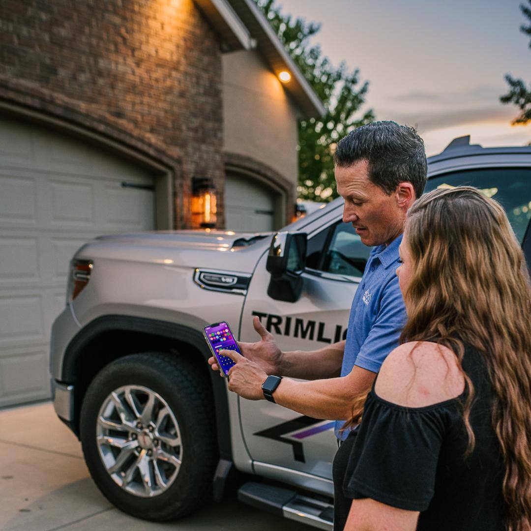 Smartphone displaying a Trimlight lighting app while a home glows with colorful exterior lights