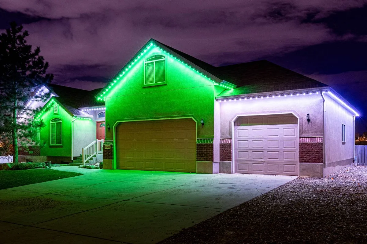 A suburban house at night features green and white LED lights along the roofline, illuminating the garage doors and front entrance. The driveway and sky are visible, with some clouds overhead.