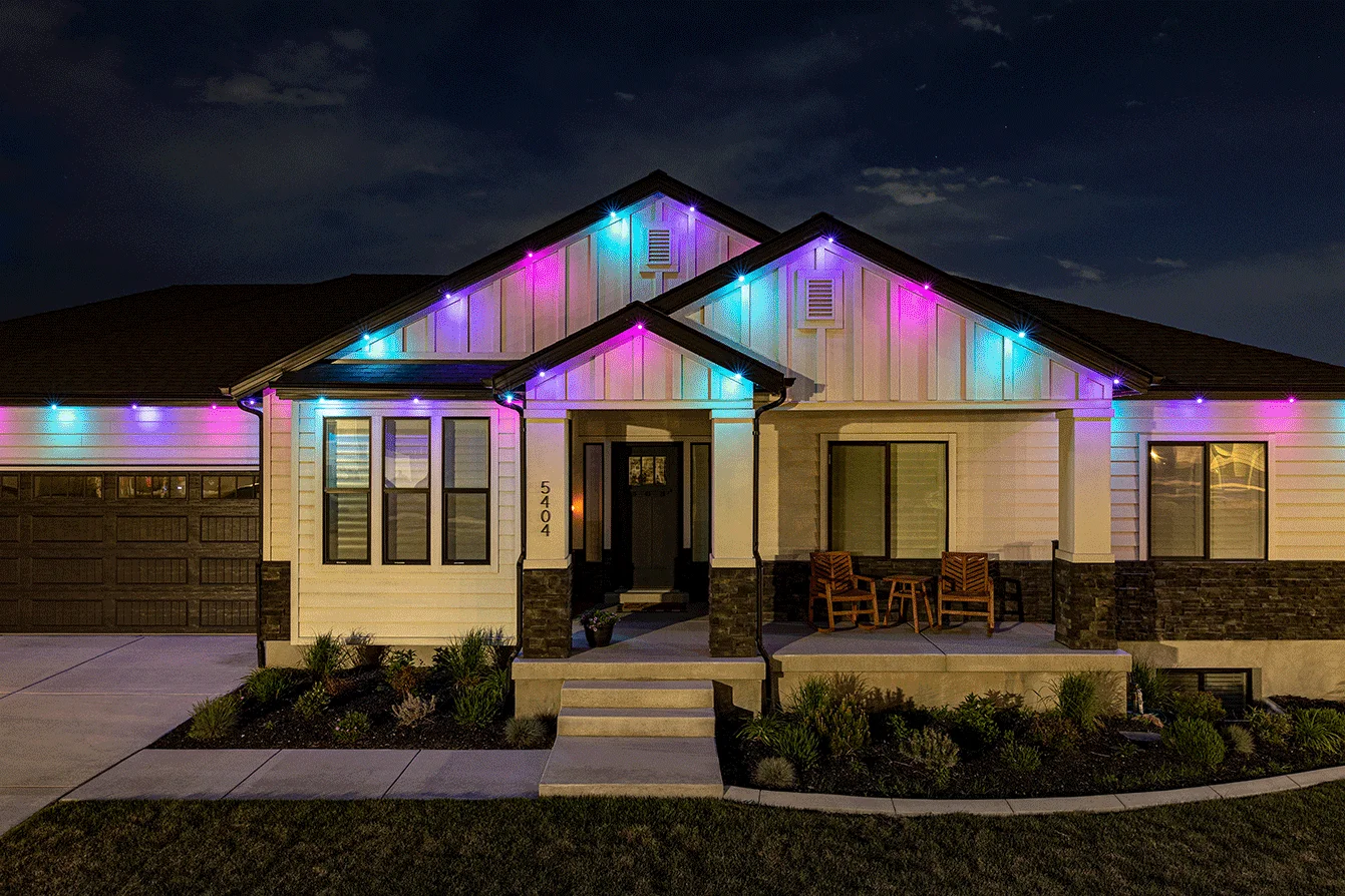 A single-story house at night displays colorful lights in blue, pink, and purple along the roofline. The porch has two wooden chairs and the yard is neatly landscaped.