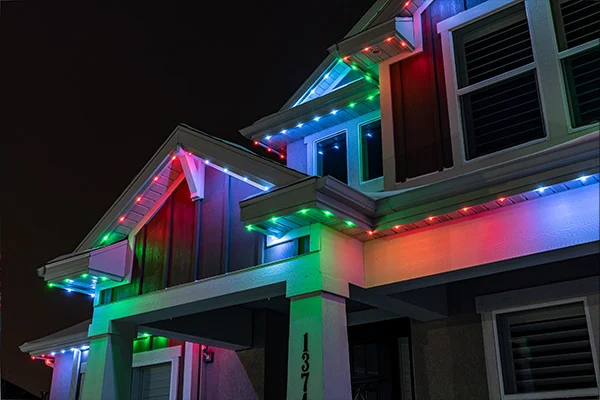 A house at night is decorated with multicolored LED lights along the roofline, displaying red, green, and blue hues. The lights illuminate the exterior and give a festive appearance.