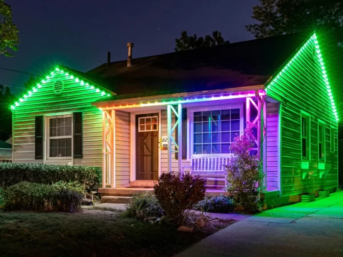 A single-story house at night with its roofline and porch illuminated by colorful LED lights in green, purple, and pink hues. Shrubs and a white bench are visible in the front yard.