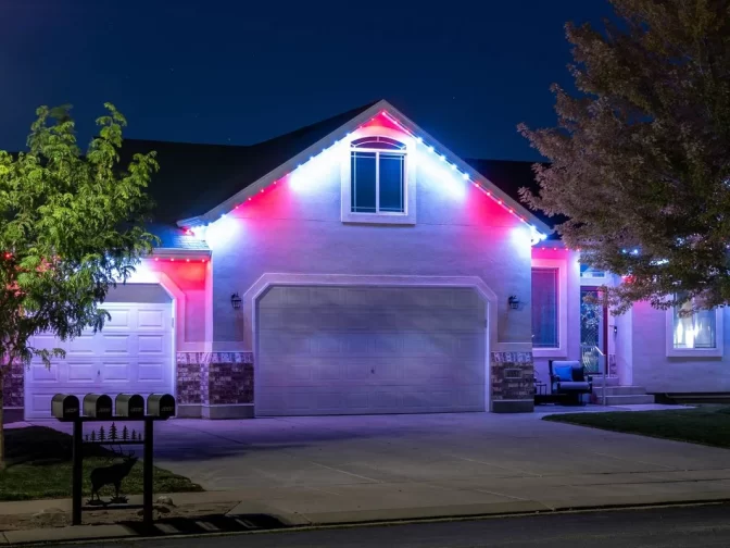A suburban house at night with white, red, and blue lights illuminating the roofline. The driveway is empty, and two trees frame the front yard. Mailboxes are visible in the foreground.