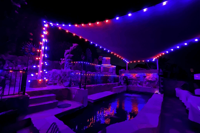 A backyard pool at night illuminated by red and blue string lights along a canopy and fence, with reflections of the lights shimmering on the water. The surrounding area appears tranquil and festive.