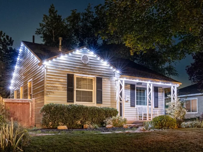 A small house with striped siding is decorated with white string lights along the roof. The front porch has a white chair, and the yard is landscaped with bushes, rocks, and grass. The photo is taken at night.