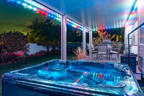 A hot tub with blue lighting sits on a covered patio at night, surrounded by outdoor furniture and multicolored string lights. The patio overlooks a yard with trees, a hammock, and a dining area.