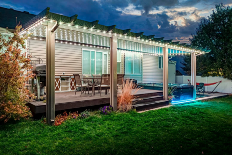 A backyard patio at dusk features a covered pergola with string lights, outdoor furniture, a grill, and a hot tub with blue lighting, surrounded by grass and landscaping.