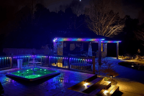 A backyard at night with a glowing hot tub, colorful LED lights along the deck and pergola, and trees in the background. The scene is reflected on the wet wooden deck, creating a vibrant, relaxing atmosphere.