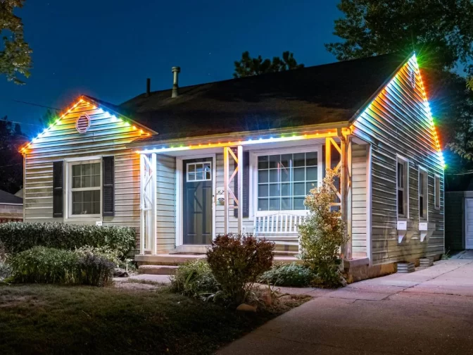 A single-story house at night with colorful LED lights outlining the roof and porch, a well-kept yard, large front windows, and a white bench on the porch.
