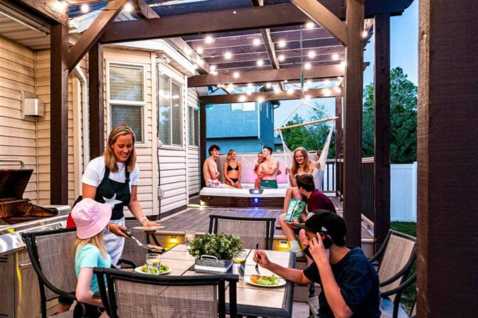 A group of people enjoy a backyard gathering; some are eating at a table under string lights while others relax in a hot tub on a raised deck, creating a lively, outdoor evening scene.