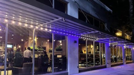 A modern restaurant with large glass windows and purple-lit awnings at night. Inside, people are seated and dining, while the sidewalk outside is empty. The interior is warmly lit with visible plants and decor.