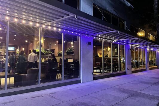A modern restaurant with large glass windows and purple-lit awnings at night. Inside, people are seated and dining, while the sidewalk outside is empty. The interior is warmly lit with visible plants and decor.