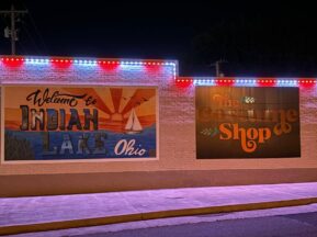 A brick building at night with two large murals: one reads Welcome to Indian Lake Ohio with a lake scene, the other says The Costume Shop in orange letters. Red, white, and blue lights outline the roof.