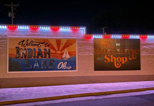 A brick building at night with two large murals: one reads Welcome to Indian Lake Ohio with a lake scene, the other says The Costume Shop in orange letters. Red, white, and blue lights outline the roof.