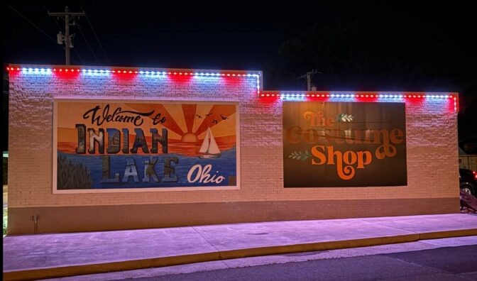 A brick building at night with two large murals: one reads Welcome to Indian Lake Ohio with a lake scene, the other says The Costume Shop in orange letters. Red, white, and blue lights outline the roof.