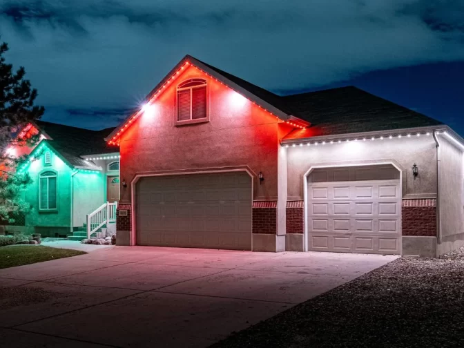 A beige house at night is decorated with red, green, and white LED lights along the roofline and garage. The driveway and front yard are visible under a cloudy sky.
