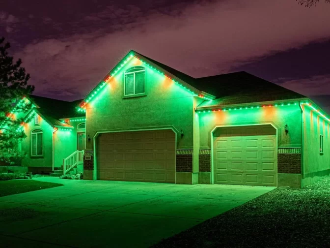 A single-story house is decorated with green and red lights along the roofline, illuminating the driveway and exterior at night under a partly cloudy sky.