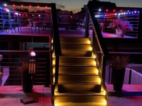 A wooden deck at dusk with illuminated stairs leading up to a second level. Both levels have red, blue, and purple string lights, potted plants, and outdoor seating, creating a cozy and inviting atmosphere.