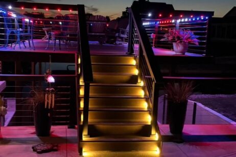A wooden deck at dusk with illuminated stairs leading up to a second level. Both levels have red, blue, and purple string lights, potted plants, and outdoor seating, creating a cozy and inviting atmosphere.