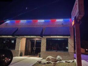 A brick building at night with blue, white, and red LED lights along the roof. Black awnings cover the windows and entrance. A wooden signpost is visible in the foreground, and rocks line the sidewalk near the doorway.