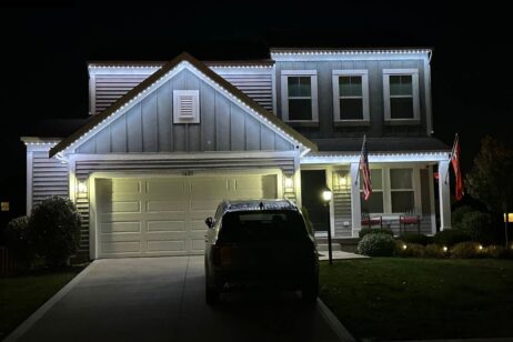 A two-story house at night is decorated with white string lights along the roofline. A car is parked in the driveway, and two flags are displayed near the front porch. The house is well-lit and surrounded by bushes.