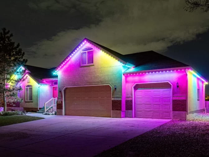A single-story house at night with its roof lined in bright, colorful LED lights glowing pink, blue, yellow, and purple, creating a vibrant and festive atmosphere.