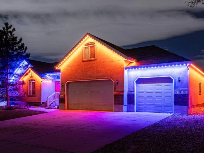 A single-story house at night is decorated with bright red, orange, and blue LED lights along the roofline, illuminating the driveway and garage with colorful glows under a cloudy sky.