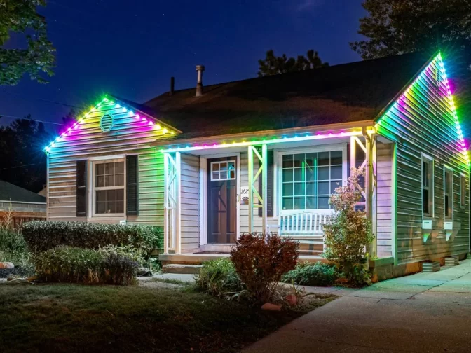 A single-story house at night with colorful LED lights outlining the roof, glowing in shades of pink, blue, yellow, and green. The house has a porch swing, shrubs, and a driveway beside it.