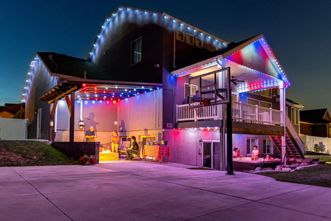 A house at dusk decorated with colorful string lights, featuring a balcony, basketball hoop, and people gathered on the patio and under the covered area in the backyard.