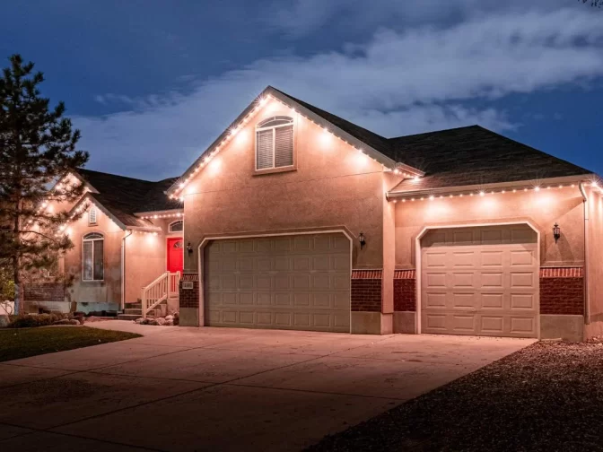 A single-story suburban house at dusk features string lights outlining the roof, a large driveway, a double garage, a single garage, and a red front door. The sky is partly cloudy with some trees visible.