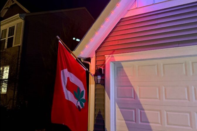 A red flag with a white O and a green leaf hangs beside a garage; red and blue lights illuminate the roofline of the house at night.