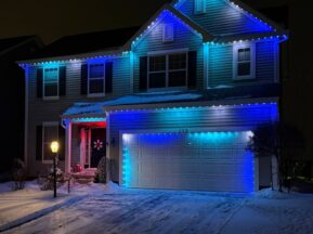 A two-story house is decorated with blue and white lights along the roofline at night, with snow covering the yard, trees, and driveway. A lit lamppost stands near the front walkway.