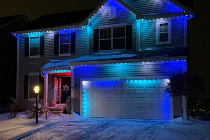 A two-story house is decorated with blue and white lights along the roofline at night, with snow covering the yard, trees, and driveway. A lit lamppost stands near the front walkway.