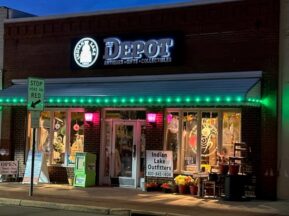 A brick storefront at night with a lit Depot Antiques • Gifts • Collectibles sign, festive window displays, green lights above the awning, and a sidewalk sign that reads Indian Lake Outfitters. A Stop Here on Red sign is nearby.