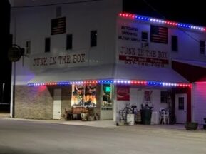 A white two-story building at night with red, white, and blue lights on the roof trim. The sign reads JUNK IN THE BOX, advertising antiques and unique military items for sale. Some vintage items are displayed outside.