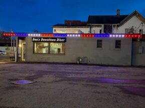 A small gray diner called Don’s Downtown Diner, with red, white, and blue lights along the roof, sits beside an empty parking lot at dusk.