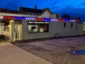 A small diner called Dons Downtown Diner is lit with red, white, and blue string lights along the roof at dusk. The building sits next to an empty parking lot with a puddle.