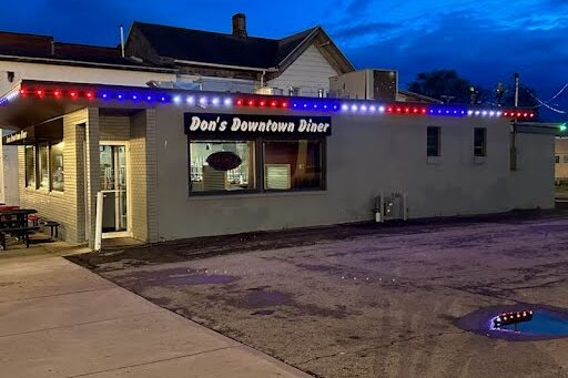 A small diner called Dons Downtown Diner is lit with red, white, and blue string lights along the roof at dusk. The building sits next to an empty parking lot with a puddle.