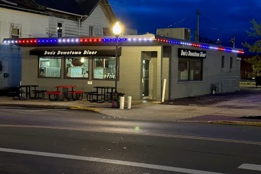 A small diner called Dons Downtown Diner is lit with red, white, and blue lights along the roof at dusk. There are tables outside, and the street in front is quiet and empty.