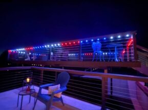 A deck at night is decorated with red, white, and blue string lights. There are metal tables and chairs on the upper level and a blue Adirondack chair with a side table and candle on the lower level.