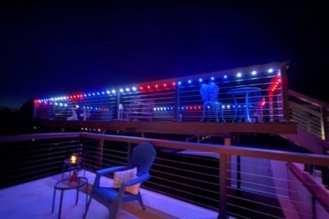 A deck at night is decorated with red, white, and blue string lights. There are metal tables and chairs on the upper level and a blue Adirondack chair with a side table and candle on the lower level.