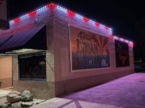A brick building at night with red, white, and blue lights along the roofline. A large mural on the wall reads Welcome to Indian Lake, Ohio. The ground is lit by outdoor lights.