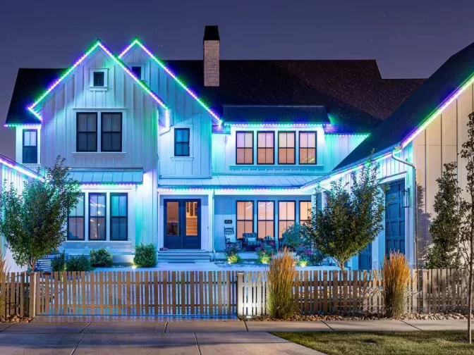 Two-story modern farmhouse with white siding, large windows, and a front porch, decorated with glowing blue and green lights along the roofline at night; fenced yard with trees and shrubs.