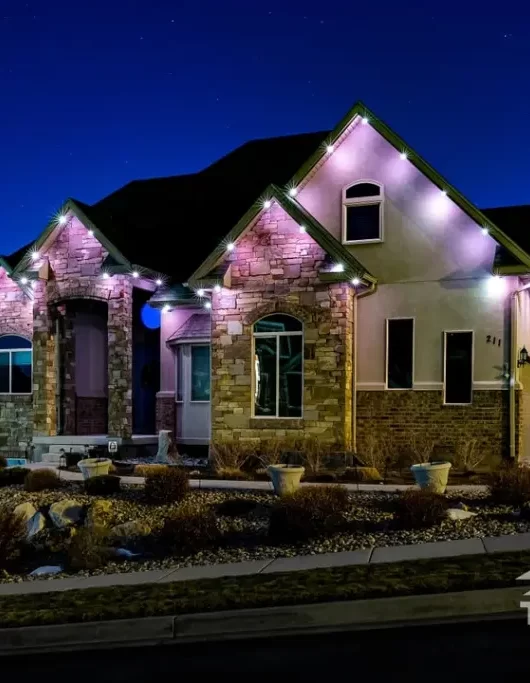 A modern house with brick and stucco exterior is illuminated by trim lights along the roofline at night. The sky is dark and clear. The Trimlight logo appears in the lower right corner.