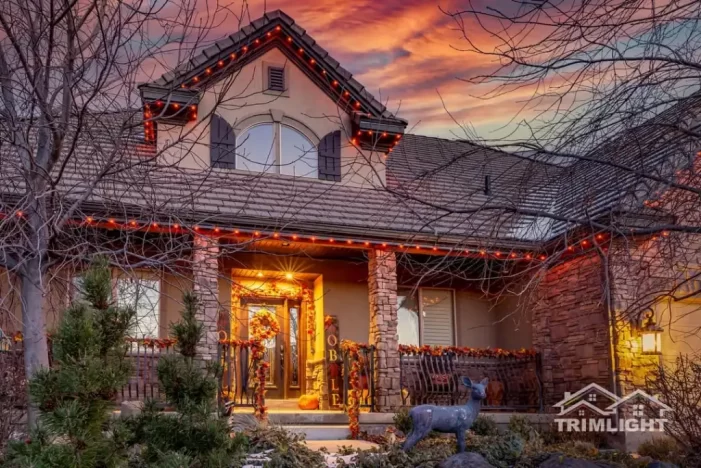 A two-story house decorated with orange and red lights and autumn garlands, with a sunset sky in the background and leafless trees in front. The porch is warmly lit and there is a deer statue in the yard.