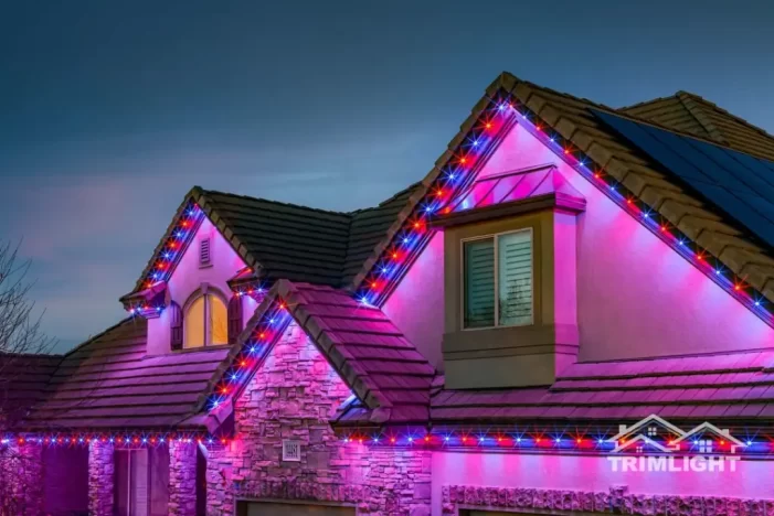 A house with stone and stucco exterior is decorated with red, blue, and white LED lights along the roof lines at dusk. The sky is darkening, and the Trimlight logo appears in the bottom right corner.