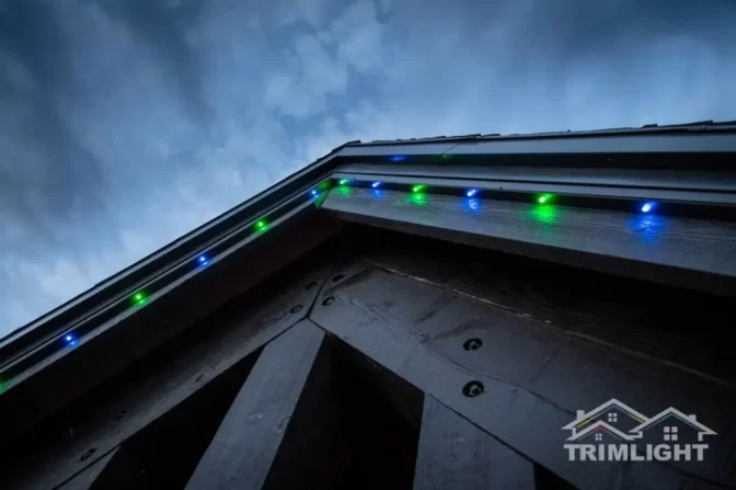 Close-up view of a house roofline at dusk with blue and green LED lights installed along the edge, illuminating the dark wooden beams below. The Trimlight logo is visible in the bottom right corner.